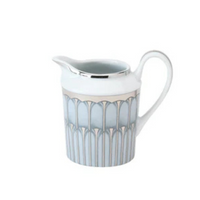 A white Limoges porcelain pitcher from Philippe Deshoulieres BIA's Arcades Platinum Dinnerware features a silver rim and handle with blue and silver geometric patterns around the body.