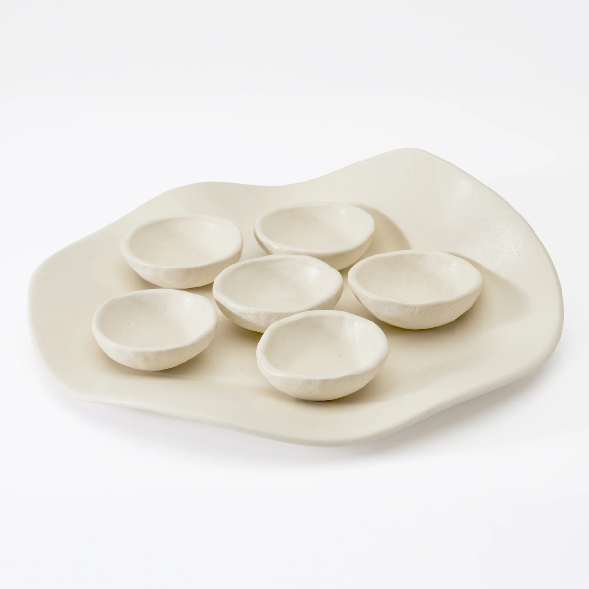 The Dumae Evie Seder Plate features an off-white, organically shaped handmade plate with six matching round ceramic bowls. Its minimalist, modern design makes it a stylish contemporary Passover Seder plate. Shown on a plain white background.