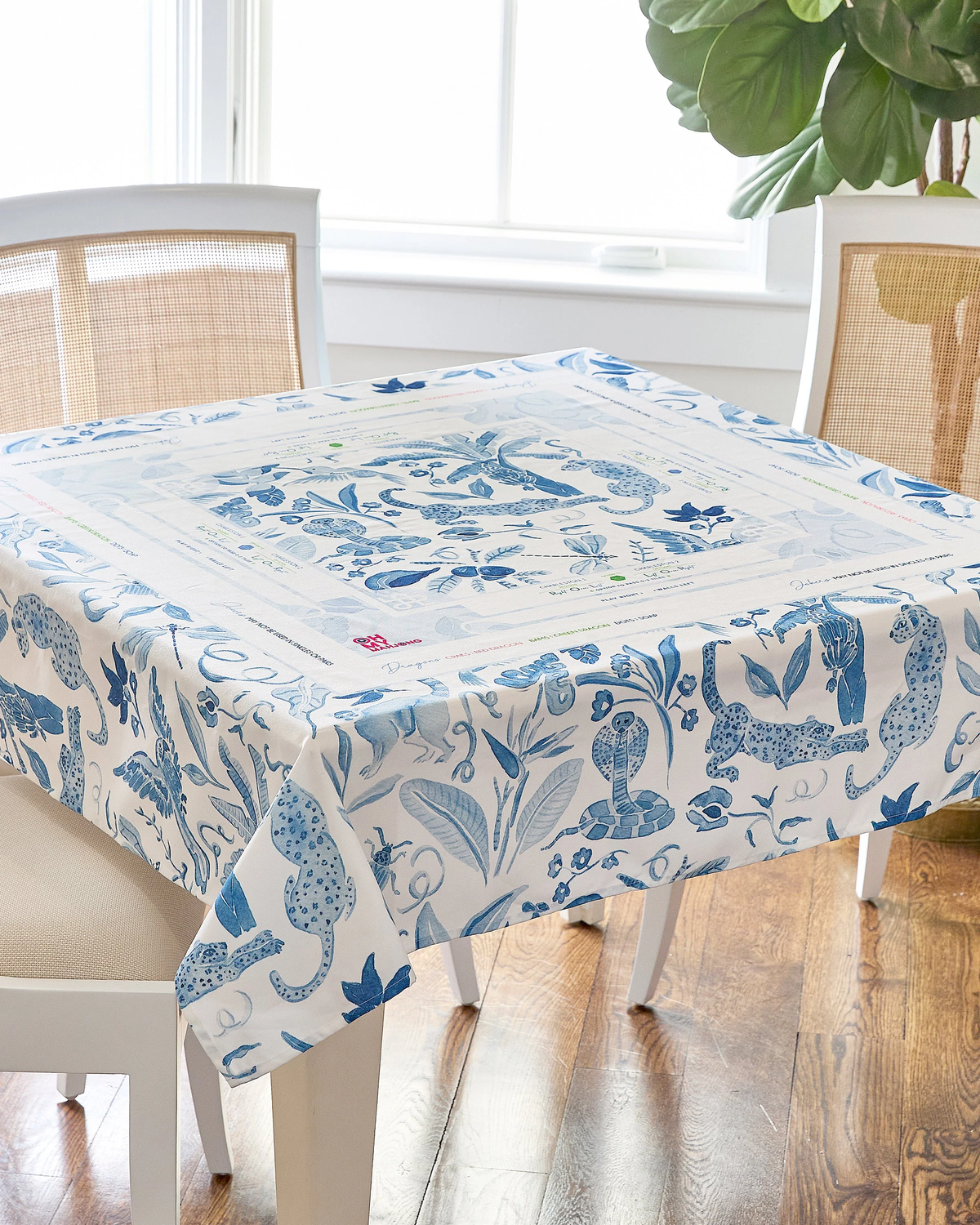 A square table covered with the Oh My Mahjong Mahjong Tablecloth, featuring a blue and white pattern of animals and plants, is surrounded by white chairs on a wooden floor. A large green plant sits near a bright window in the background.