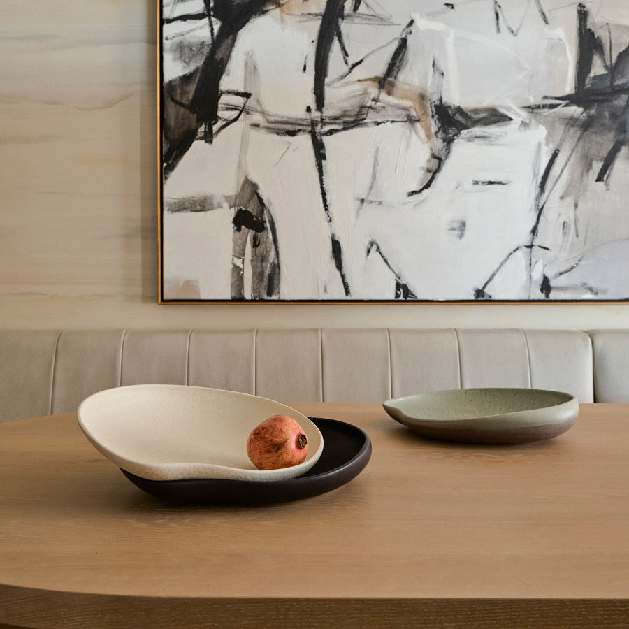 A modern dining table features two minimalist Greg Natale Tefra Bowls—one textured and filled with a pomegranate. An abstract black-and-white painting and a cushioned beige bench complete the stylish interior.