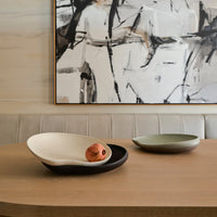 A modern dining table features two minimalist Greg Natale Tefra Bowls—one textured and filled with a pomegranate. An abstract black-and-white painting and a cushioned beige bench complete the stylish interior.