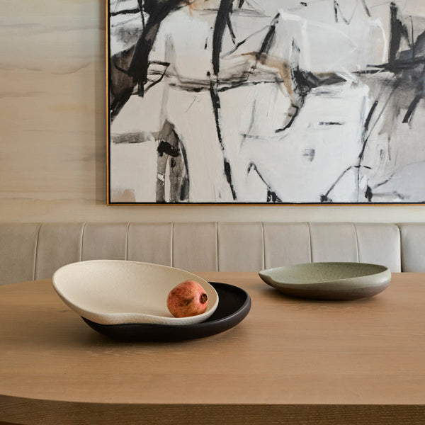 A modern dining table features two minimalist Greg Natale Tefra Bowls—one textured and filled with a pomegranate. An abstract black-and-white painting and a cushioned beige bench complete the stylish interior.