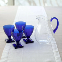 The Caspari Acrylic Handled Pitcher in clear and blue sits on a white runner beside four square-based blue goblets, all arranged together on a light-colored table.