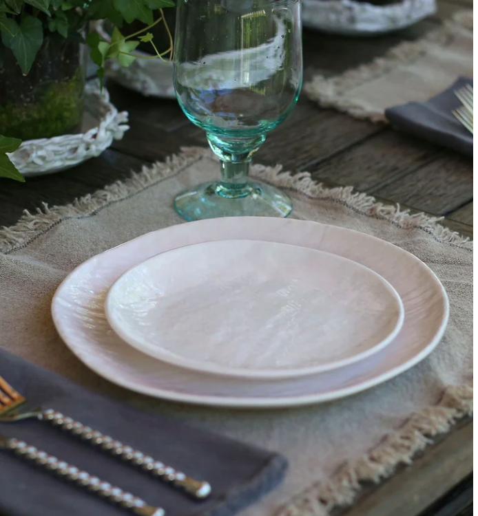 A rustic table setting with Relish Weave Melamine Dinnerware in white, a clear water glass, dark-handled silverware, and a fringed beige placemat on wood, set against greenery.
