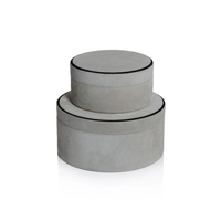 Two round Aldo Suede Box Grey decorative boxes by Zodax are stacked, with the smaller on top. Both feature light gray suede leather and black-trimmed edges, creating a minimalist storage set against a white background.