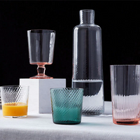 Lando Pleat Glassware by MATCH—featuring a carafe, colored tumblers, and a stemmed glass inspired by antique Italian designs—is arranged on a white surface with one glass holding orange juice against a dark background.