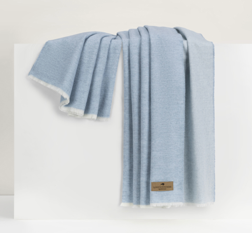 A light blue Aspen Reversible Throw by Lands Downunder with frayed edges is draped over a white surface. A small brown logo tag adds elegance to the soft, cozy blanket.