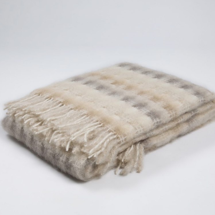 The Mia Throw by Mantas Ezcaray is a folded, soft woolen blanket featuring a beige and gray plaid pattern with fringe on one edge, highlighting artisanal quality against a plain white background.