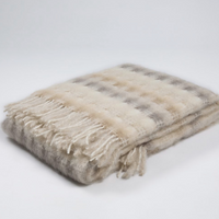 The Mia Throw by Mantas Ezcaray is a folded, soft woolen blanket featuring a beige and gray plaid pattern with fringe on one edge, highlighting artisanal quality against a plain white background.
