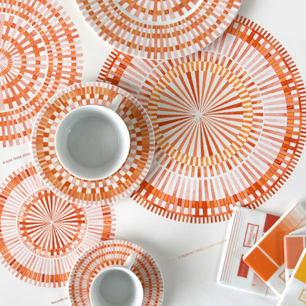Overhead view of Bernardaud Terra Rosa Dinnerware—coffee cups, saucers, plates, and placemats with orange and red geometric circles on a white surface. Includes color swatches. Made in Limoges, France.