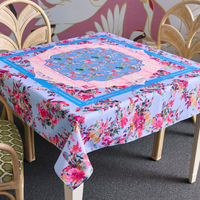A square table covered with an Oh My Mahjong Mahjong Tablecloth sits between two wicker chairs. The tablecloth features pink, blue, and red flowers arranged in decorative patterns.