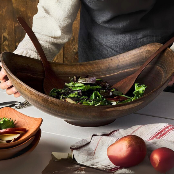 Wearing an apron, a person tosses salad in an Andrew Pearce Live Edge Wood Bowl Walnut using wooden utensils. Nearby: potato, apple slices, a striped towel, and cutting boards rest on the white table.