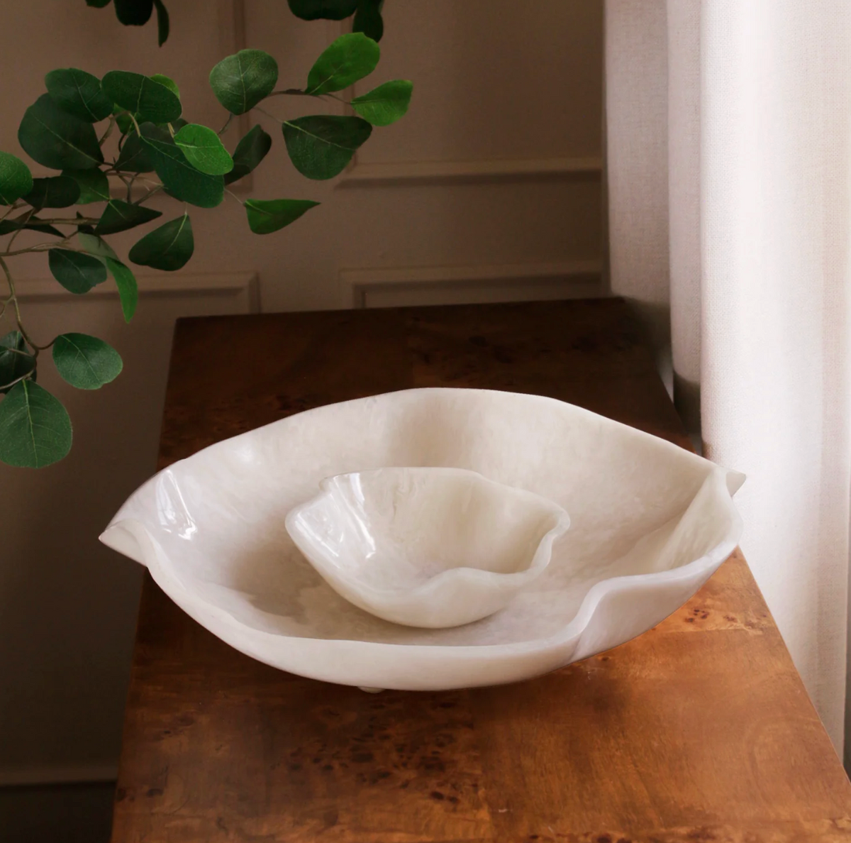 The Beatriz Ball Resin Alabaster Chip & Dip—two white, wavy-edged bowls—are stacked on a wooden table by a window with white curtains, with green leaves partially framing the scene.