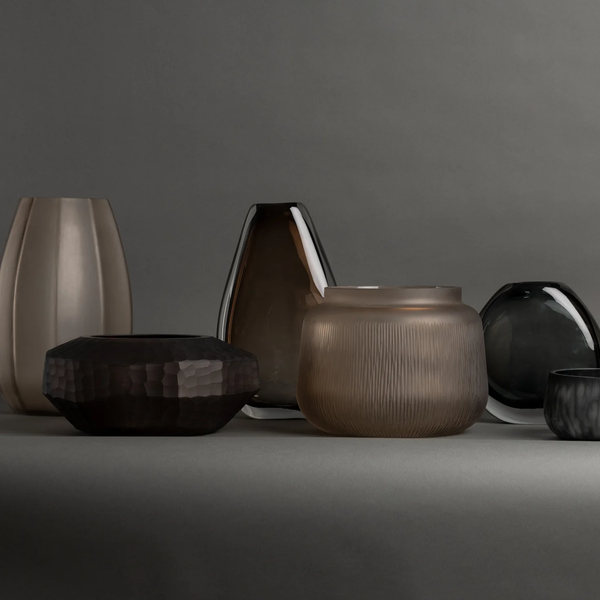 The Nellie Vase by FlowDecor Inc.—a modern, uniquely shaped vase in smokey gray—stands out on a matching gray surface and background, making it an ideal piece for stylish home decor.