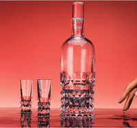The Baccarat Louxor Vodka Set of 2, featuring a diamond-cut crystal decanter and two matching shot glasses, is displayed on a reflective surface with a red backdrop as a hand reaches from the right.