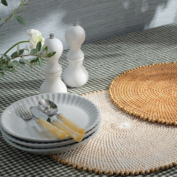 A stack of white plates with the Blue Pheasant Lulu Flatware 5 Piece Setting in Ivory sits beside two woven placemats, a vase of white flowers, and white salt and pepper shakers on a checkered tablecloth.