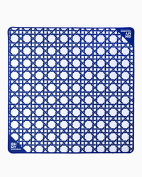 The Oh My Mahjong Travel Mini Mahjong Game Mat is a 22” x 22” square blue and white mat with a geometric pattern of interlocking lines and circles. Its corners feature small circular logos reading “OH! MY MAHJONG” and “MAHJONG.”.