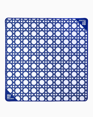 The Oh My Mahjong Travel Mini Mahjong Game Mat is a 22” x 22” square blue and white mat with a geometric pattern of interlocking lines and circles. Its corners feature small circular logos reading “OH! MY MAHJONG” and “MAHJONG.”.