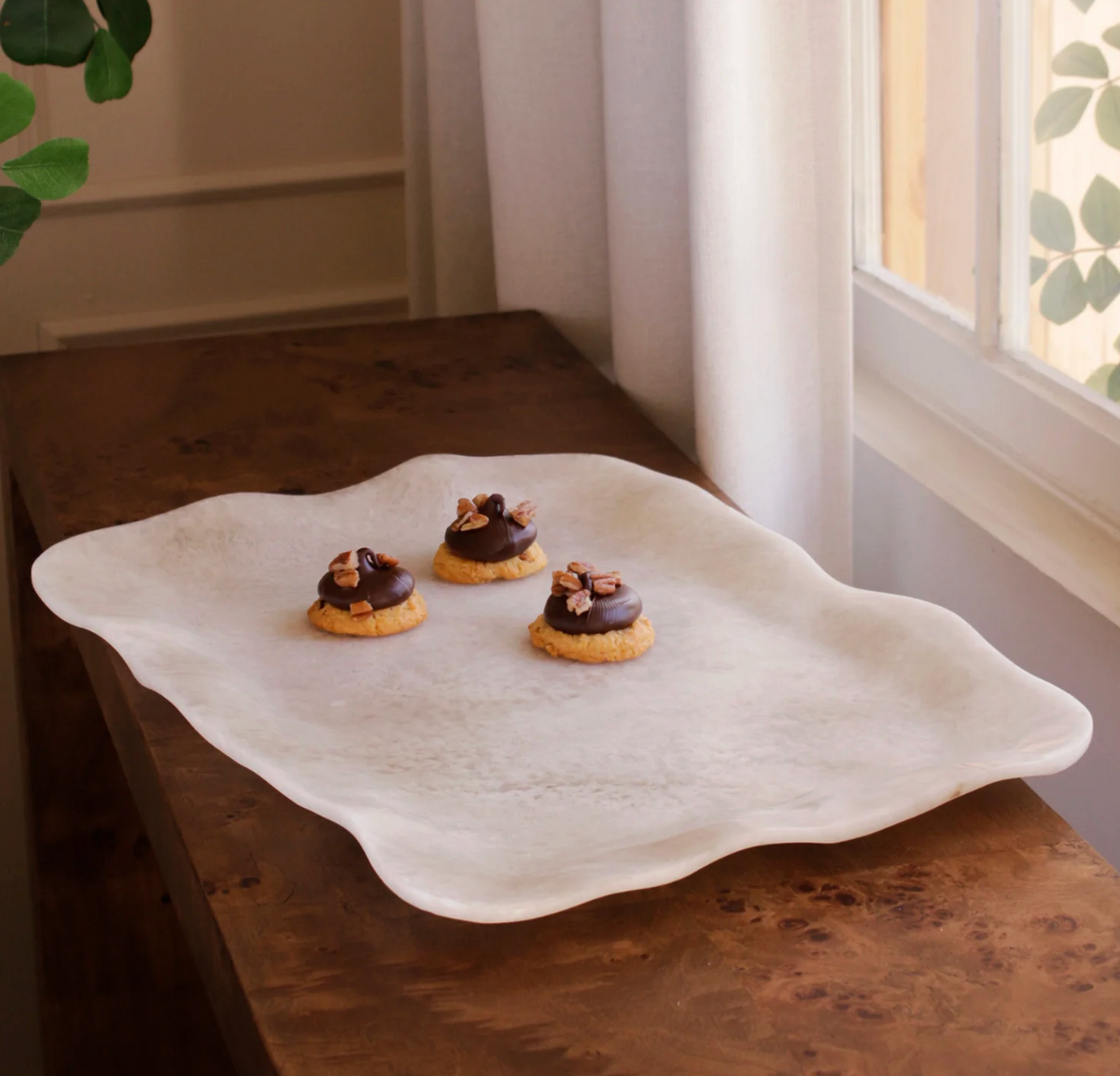 The Beatriz Ball Resin Tray Alabaster Extra Large with wavy edges rests on a wooden table by a window, holding three small cookies topped with chocolate and pecans.