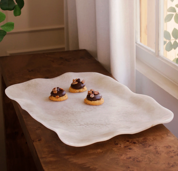 The Beatriz Ball Resin Tray Alabaster Extra Large with wavy edges rests on a wooden table by a window, holding three small cookies topped with chocolate and pecans.