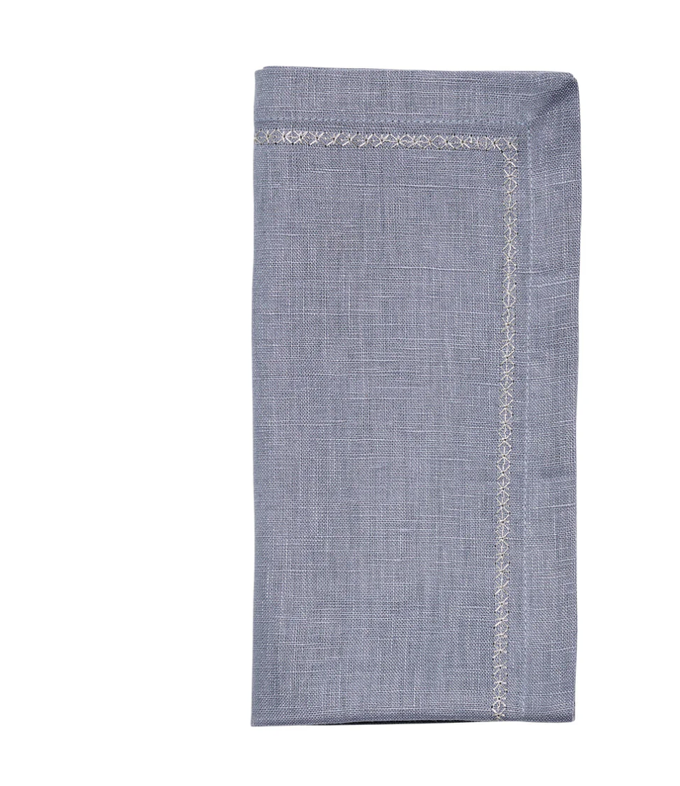 The Kim Seybert Classic Napkin - Dusty Blues (Set of 4) features a folded light blue cloth with a decorative stitched border, shown against a white background.