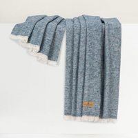 A Lands Downunder Cinder Mist Throw—with its soft blue hue, subtle textured pattern, white fringe trim, and a brown label—drapes neatly over a white surface.