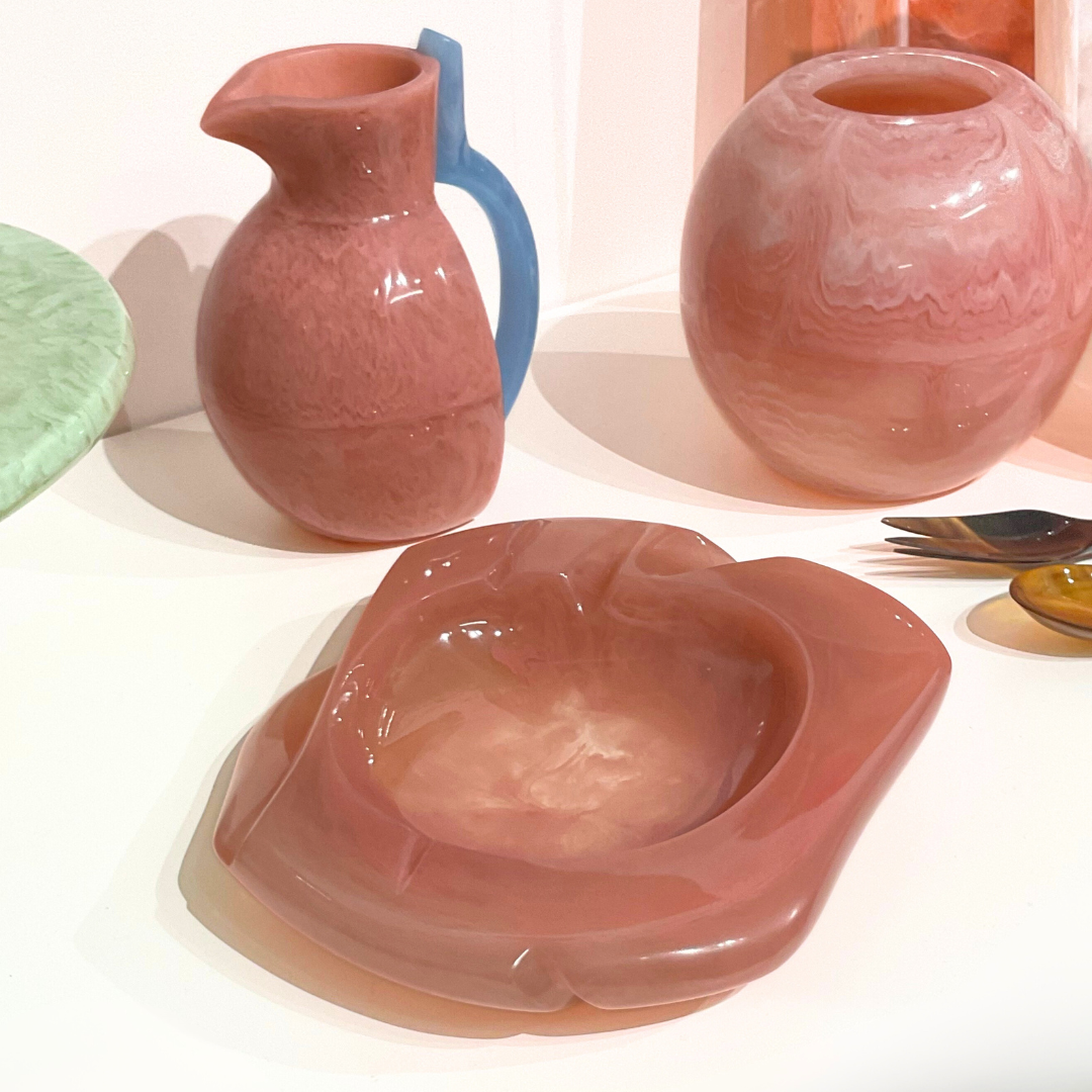 A pink glass pitcher with a blue handle, a round marbled pink vase, and the sculptural Malibu Resin Dish by Lily Juliet are displayed on a white surface alongside a durable resin bowl and other tableware.