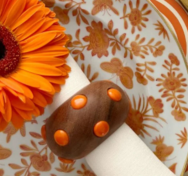 An orange gerbera daisy sits next to a Von Gern Home Candy Dots Napkin Ring Set of 4 with orange dots on a patterned plate.