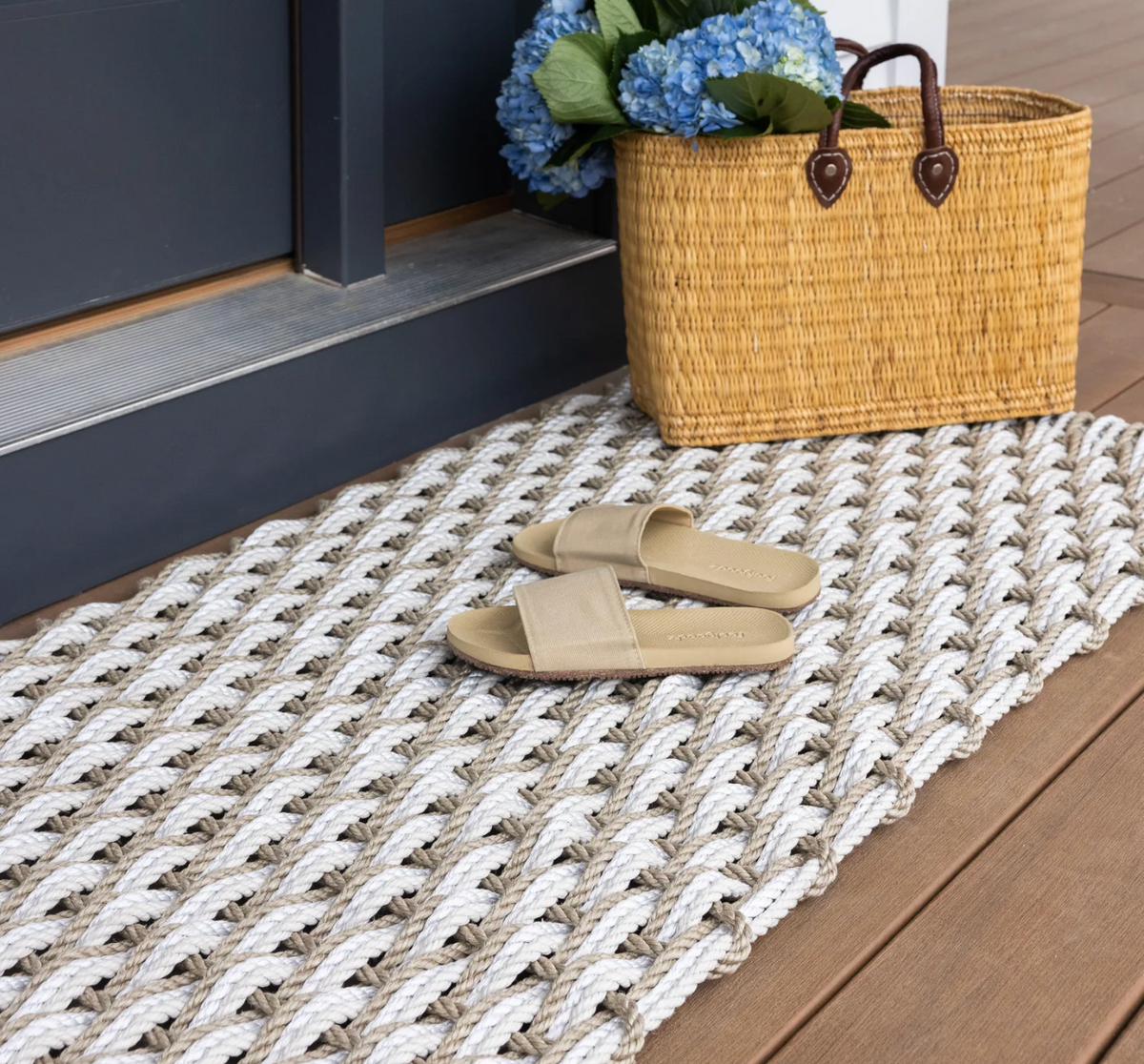 A Coastal Woven Outdoor Floormat in sand/oyster by The Rope Co (24 x 38) sits by the door. Beige slide sandals rest on the handwoven rope mat, beside a basket of blue hydrangeas on the wooden porch.
