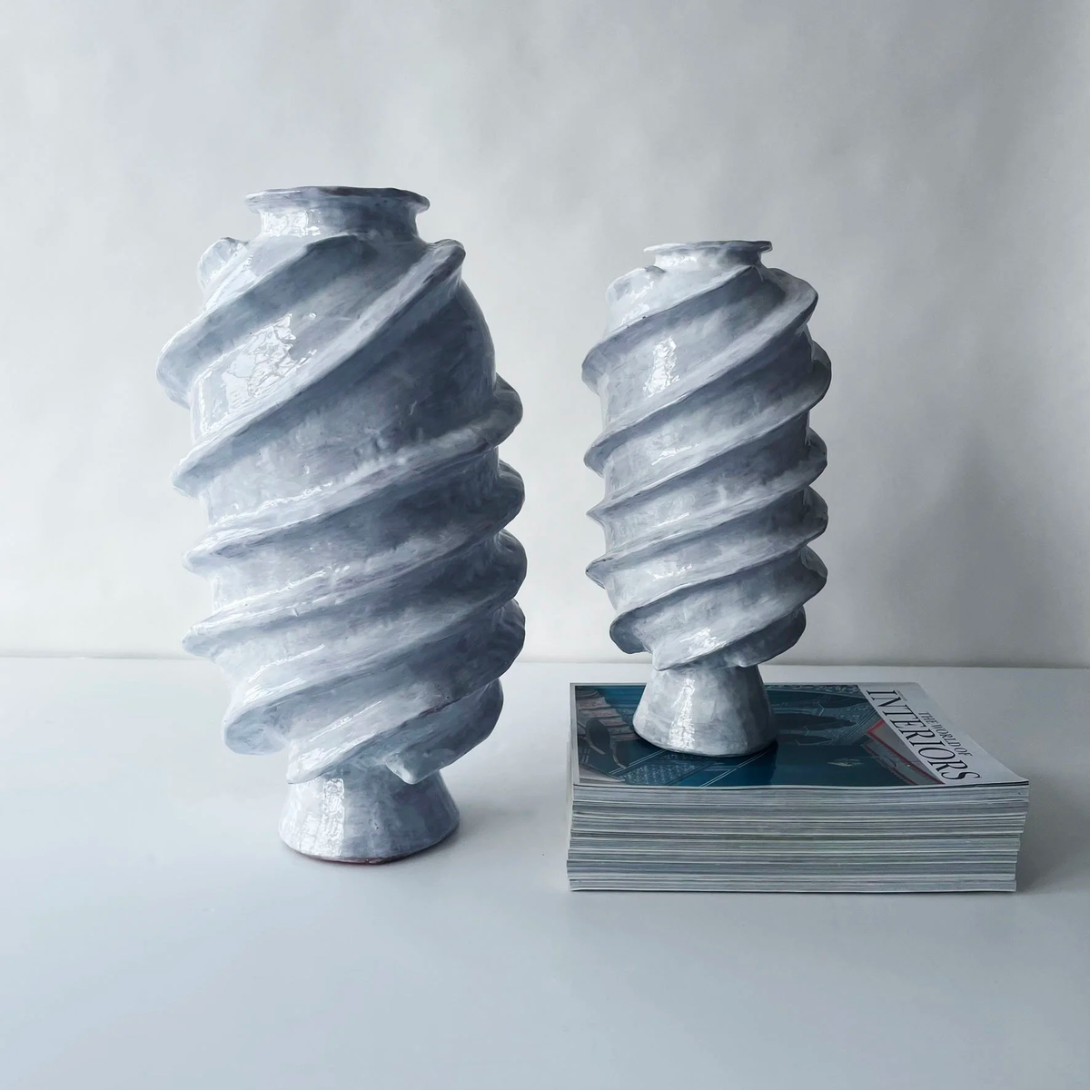Two Ithaka Vases by Ellen Robinson Studio, featuring spiral ridges and a glossy gray finish, sit on a white surface; the taller vase stands beside the shorter one, which is placed atop a stack of magazines.