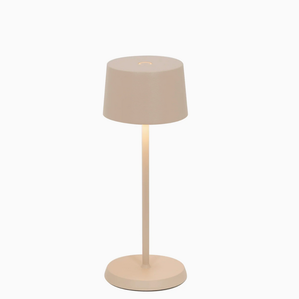The Zafferano Olivia Pro Micro Table Lamp in Sand has a minimalist design with a beige cylindrical shade, slim straight stand, and round base, all matching. Shown switched on against a white background, it emits warm downward light.