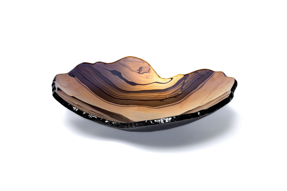 The Mapa Bowl - Bronze by Gardeco is a shallow, bowl-shaped glass dish featuring layered brown, amber, and black tones reminiscent of natural rock or agate patterns, making it a luxurious accent piece when displayed against a white background.