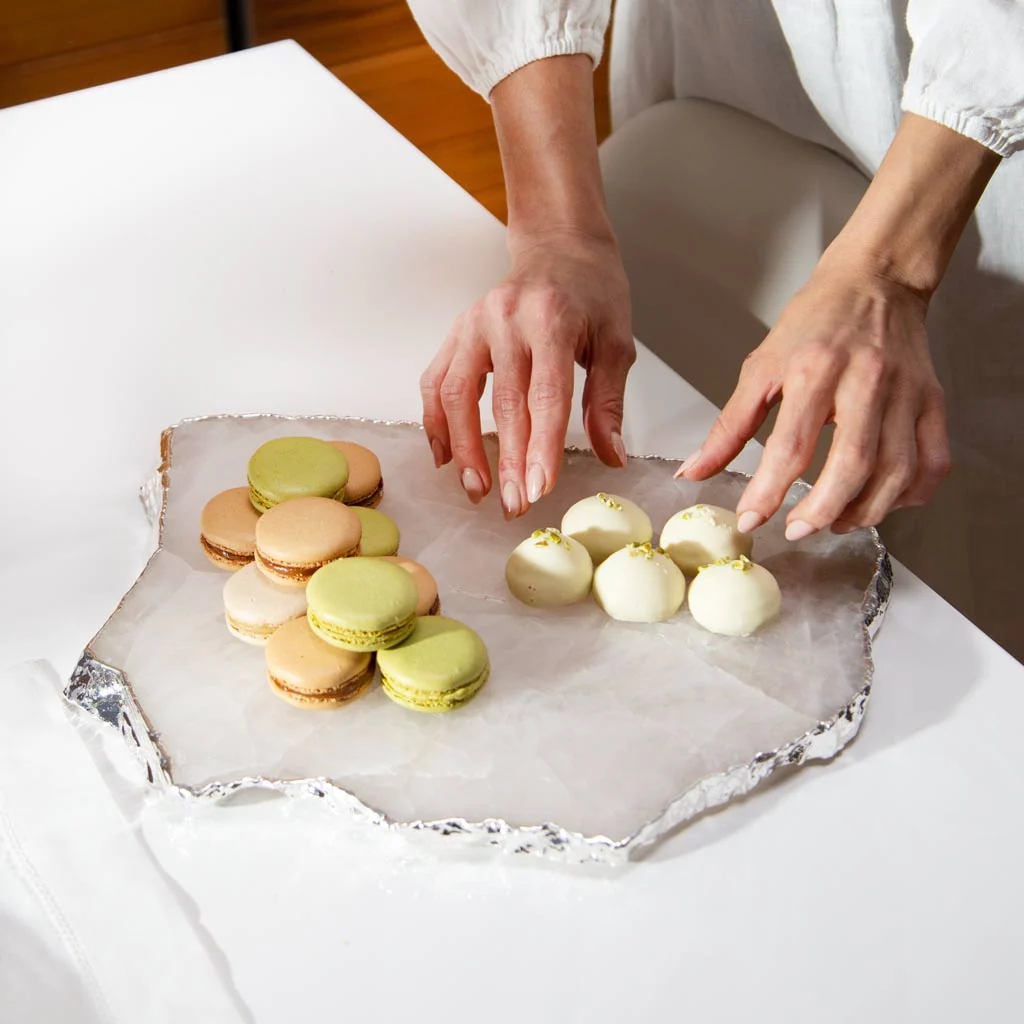A person arranges green and beige macarons and white round desserts on the Anna New York Kiva Large Platter in Crystal/Silver, lined with parchment paper and foil.