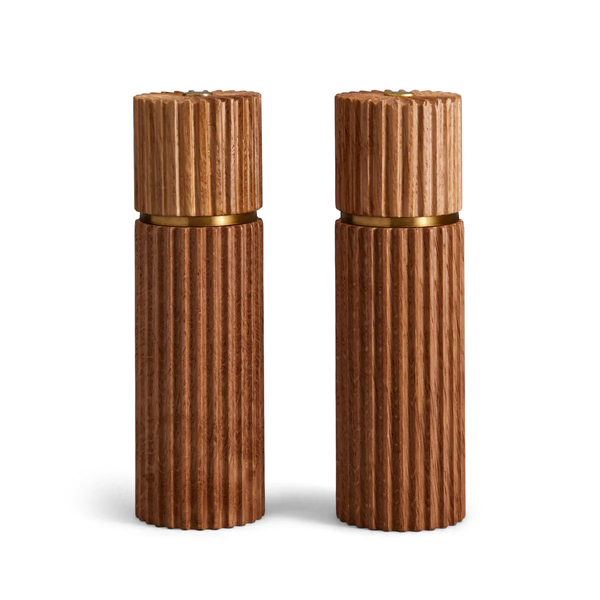 The Ionic Salt & Pepper Mill Set by L'Objet features two matching cylindrical mills in natural European oak with vertical grooves and gold accents, displayed side by side against a white background.