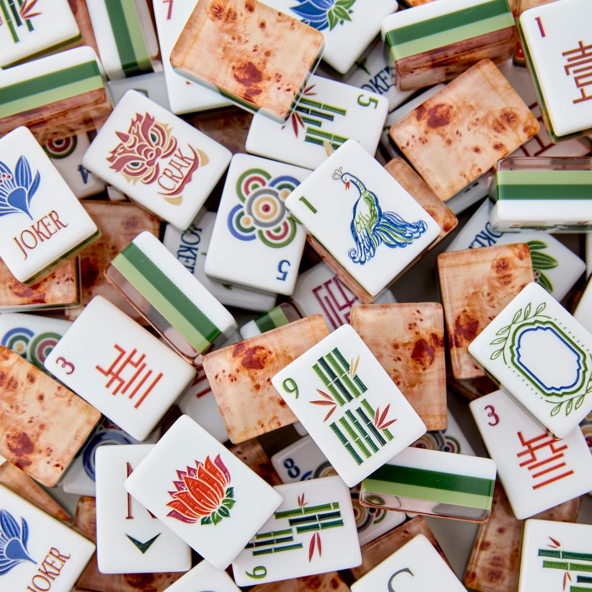A vibrant assortment of the Mahjong Tiles Set - Heritage by Oh My Mahjong, featuring an heirloom-quality finish and adorned with a variety of symbols, numbers, flowers, birds, and Jokers, is artfully arranged in a random overlapping display.