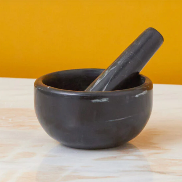 The Be Home Sicily Black Marble Mortar & Pestle rests on a white marble surface with an orange-yellow backdrop—ideal for those who appreciate stylish marble kitchenware for grinding spices.