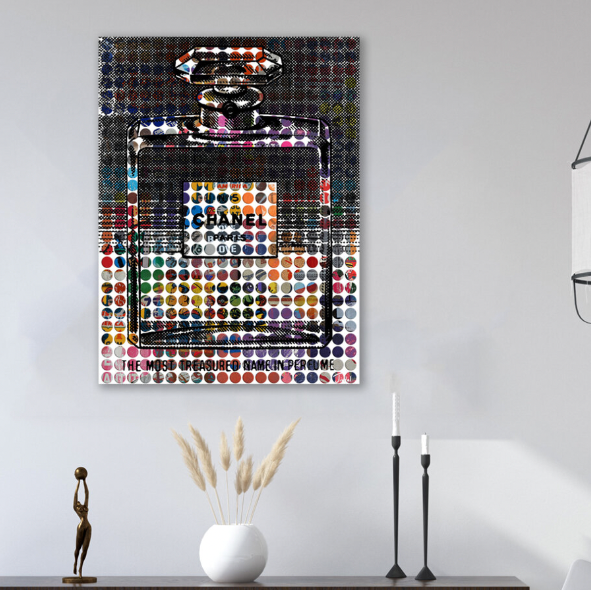Vintage Chanel Art by Sabeena Jindal, featuring a colorful Chanel No. 5 perfume bottle with circular patterns, hangs above minimalist decor—a white vase with dried grass and two candlesticks—on a white wall.