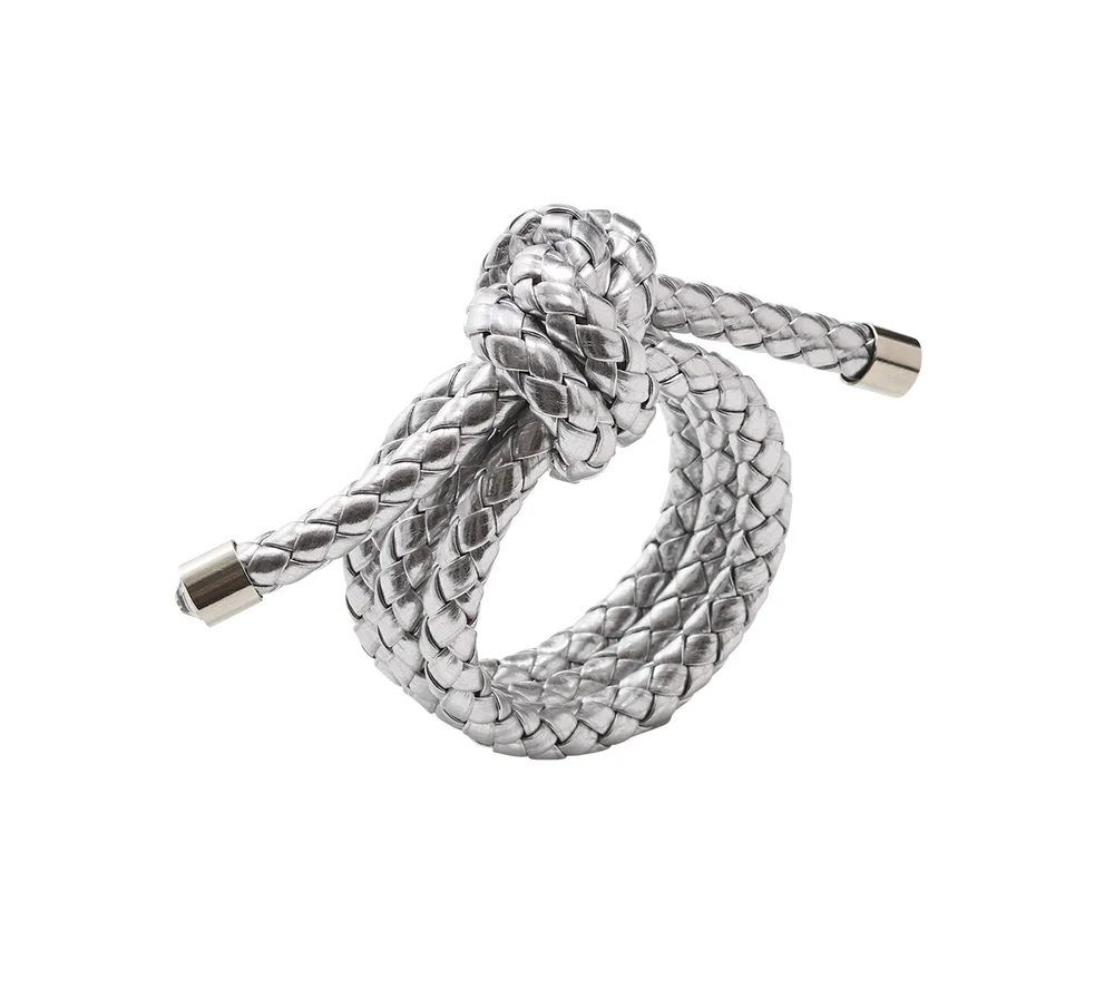 The Rope Knot Napkin Ring Set of 4 by Kim Seybert features metallic silver braided cords with metal-capped ends, intricately knotted in a circular design inspired by coastal style, shown on a plain white background.