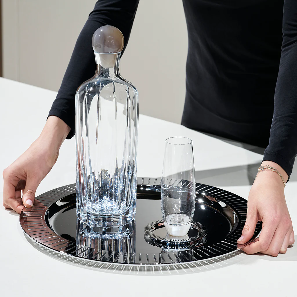 A person in a black long-sleeve shirt holds the Anna New York Striations Silver Round Serving Tray, featuring a tall crystal decanter, matching glass, and a small candle on a white countertop.