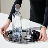 A person in a black long-sleeve shirt holds the Anna New York Striations Silver Round Serving Tray, featuring a tall crystal decanter, matching glass, and a small candle on a white countertop.