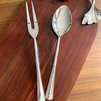 The Mary Jurek Alta Meat Serving Fork, with its hammered handle design, sits on a wooden cutting board beside a decorative silver leaf dish—an elegant touch for any serveware collection.