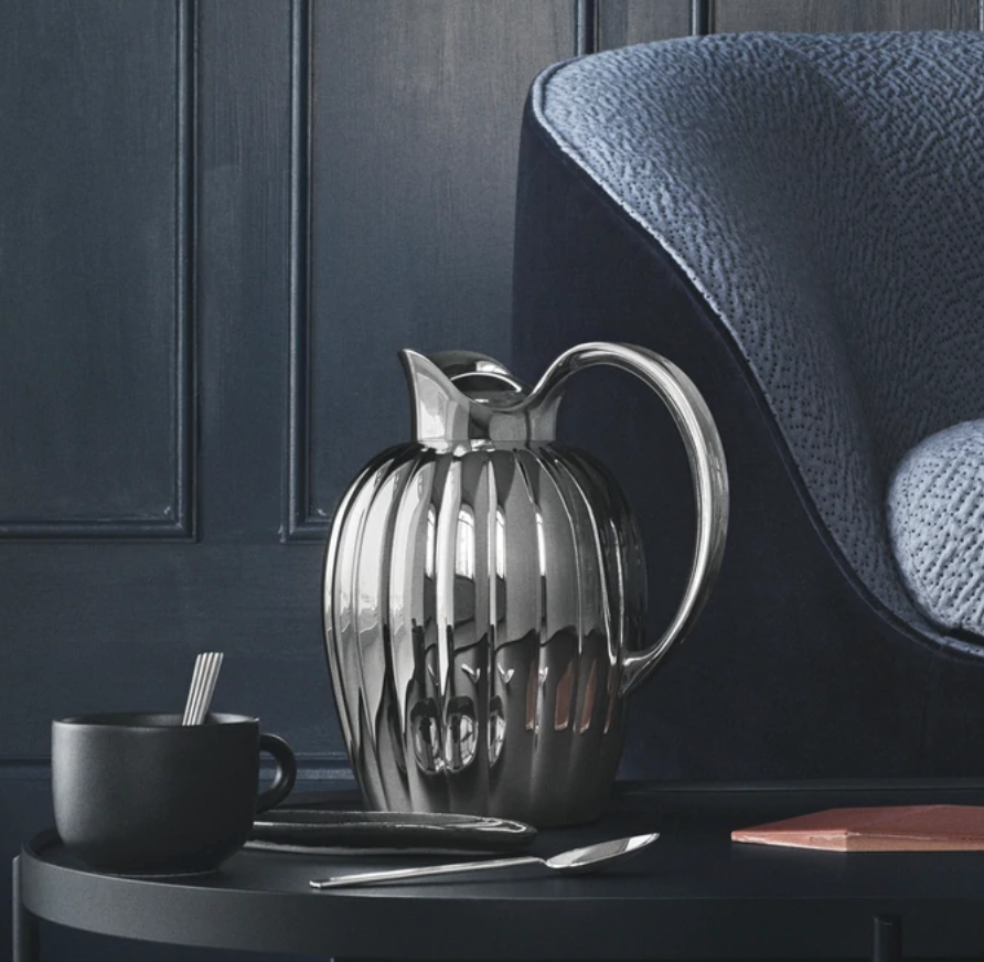 A Bernadotte New Thermo Jug w/Push Stopper by Georg Jensen sits on a round black table beside a matte black cup with spoons, a butter knife, and a closed book, set against a dark blue wall and textured blue armchair.