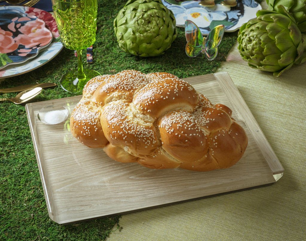 A braided challah loaf topped with sesame seeds sits on the Apeloig Solid Acrylic Challah Board & Knife, surrounded by floral plates, artichokes, and a green glass on a grass-like tablecloth.