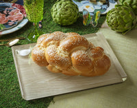 A braided challah loaf topped with sesame seeds sits on the Apeloig Solid Acrylic Challah Board & Knife, surrounded by floral plates, artichokes, and a green glass on a grass-like tablecloth.