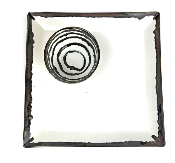 The Michael Wainwright Linea Dip Bowl, featuring black and white concentric circles, rests on a large square plate with a dark border and white center, both set against a white background.