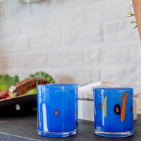 Two Abigails Torcello Stripes Cobalt DOF mouth-blown glass tumblers with colorful accents sit on a dark textured surface, with a wicker basket and green leaves blurred in the background.