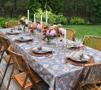 An outdoor table set for a formal meal features the stunning Kim Seybert Paisley Vine Tablecloth in Blue & Gray (54x126″), accented with a floral centerpiece, tall white candles, crystal glasses, patterned plates, napkins, and bamboo chairs on a wooden deck surrounded by a lush green garden.