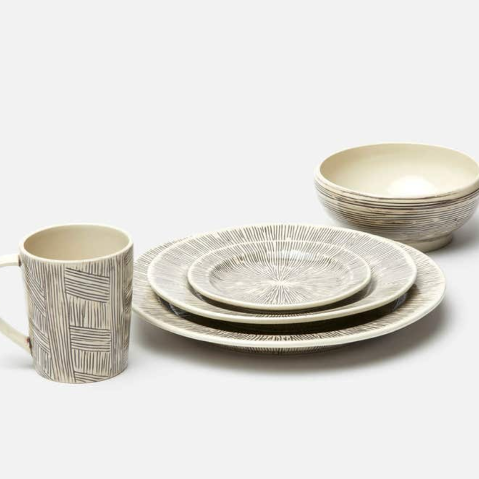 The Nolan Dinnerware Black Cream set by Blue Pheasant includes a mug, two bowls, and three plates with cream bases and linear gray patterns, offering artisanal charm to your tabletop. Each ceramic piece highlights unique handpainted details.