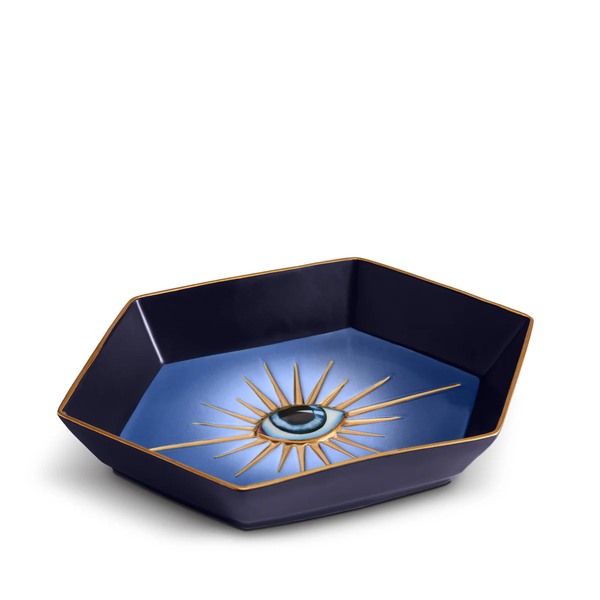 The L’Objet Lito Eye Hexagon Tray features navy blue porcelain with 24-karat gold trim, a central eye motif, and radiating gold lines on a gradient blue background.