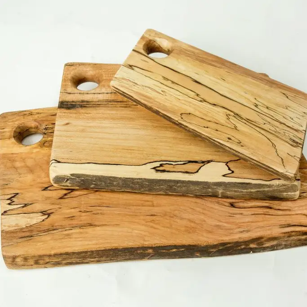 The Peterman Spalted Maple Rectangle Cutting Board 18" is an artisan board with a round corner hole, shown stacked with other wooden boards of varying sizes and finishes against a plain white background.